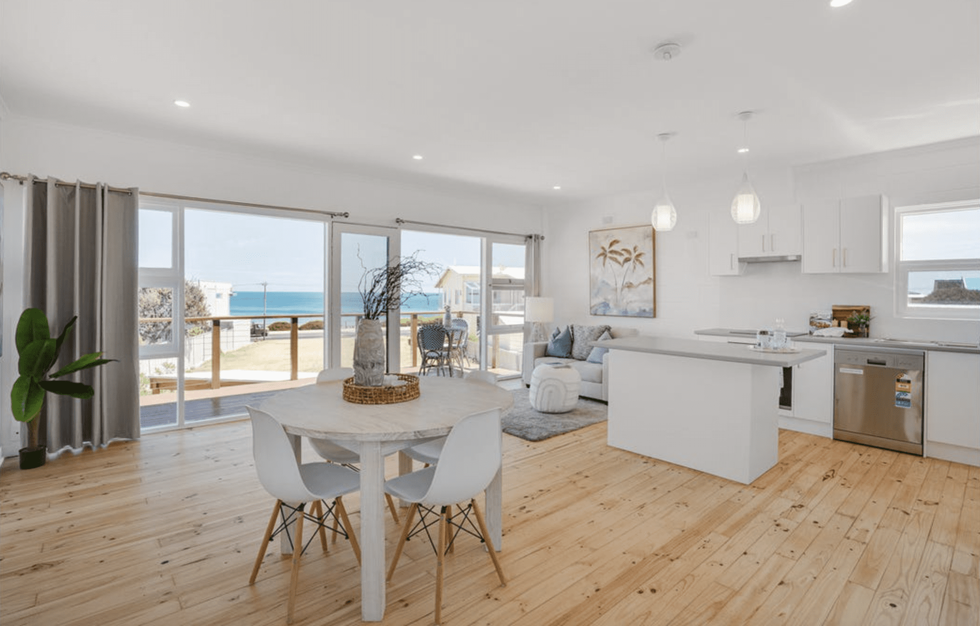 Interior view of a property showcasing a stunning ocean view from the living area, featuring large glass doors and windows opening to a wooden deck.
