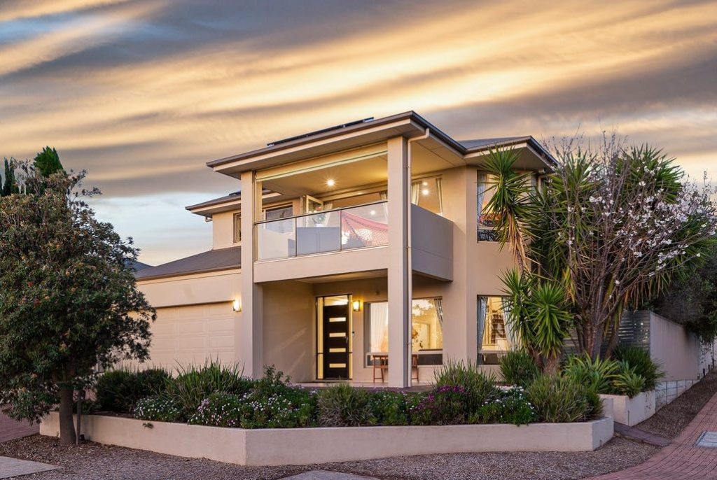 A stunning modern two-storey home with a spacious balcony, elegant lighting, and lush landscaping, captured during a picturesque sunset.