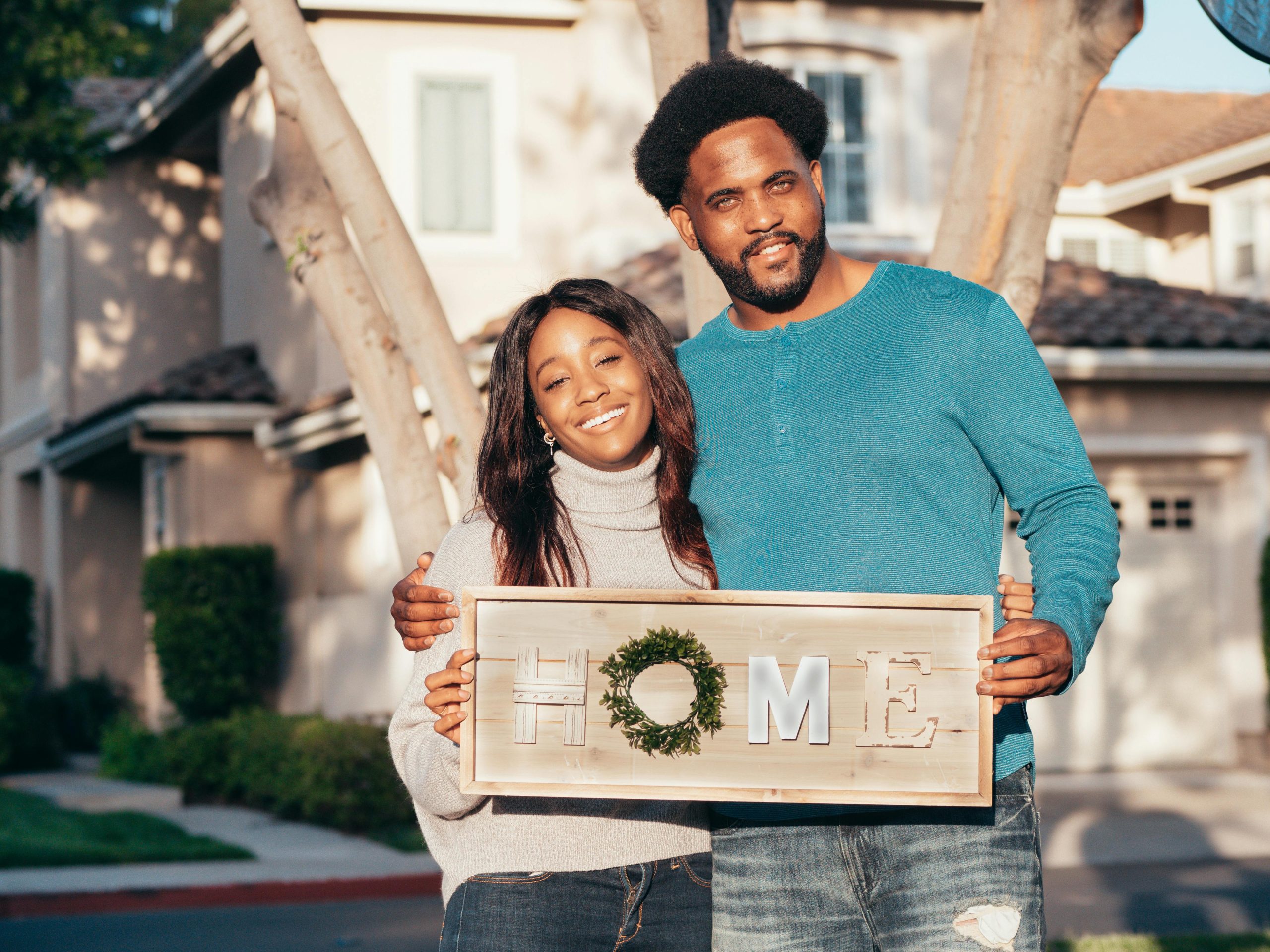 A happy couple standing outdoors, holding a wooden "HOME" sign, symbolising the joy of owning a home and achieving property ownership goals.