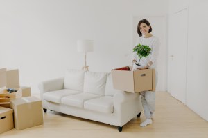 A cheerful woman standing in a bright living room, holding a cardboard box with a potted plant, symbolising a fresh start and moving into a new home.