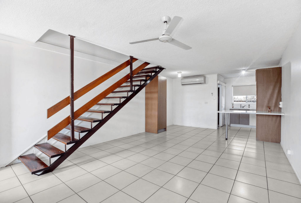 Modern open-plan living area of a Mackay townhouse featuring tiled floors, a timber staircase with open risers, ceiling fan, and split-system air conditioning leading into a compact kitchen.