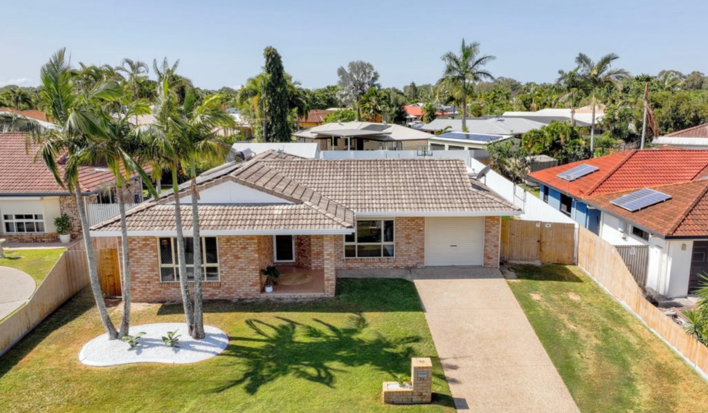 Brick single-storey home on a 700m² block in Bucasia, QLD, featuring a tiled roof, palm trees, manicured lawn and a wide driveway leading to a single garage.